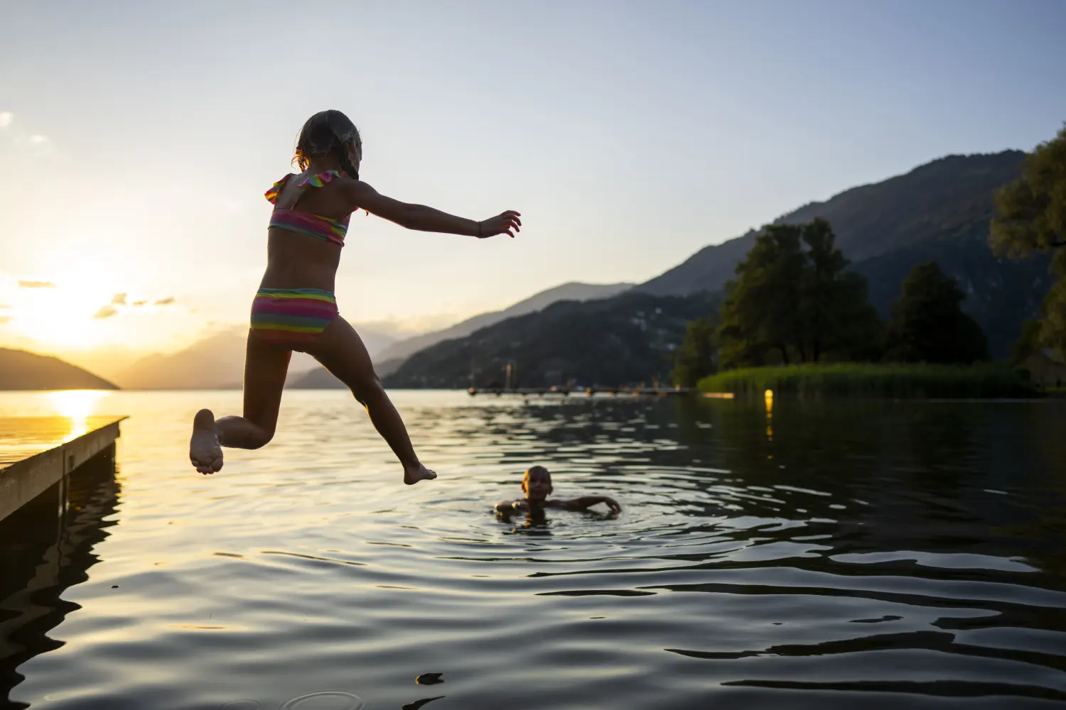 Ein Mädchen springt bei Sonnenuntergang von einem Holzsteg in den warmen Millstätter See