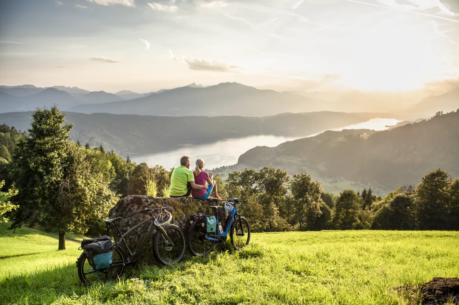 Ein paar sitzt nach ihrer Radtour auf einem Berg auf einen großen Stein und schaut bei Sonnenuntergang auf den See hinab